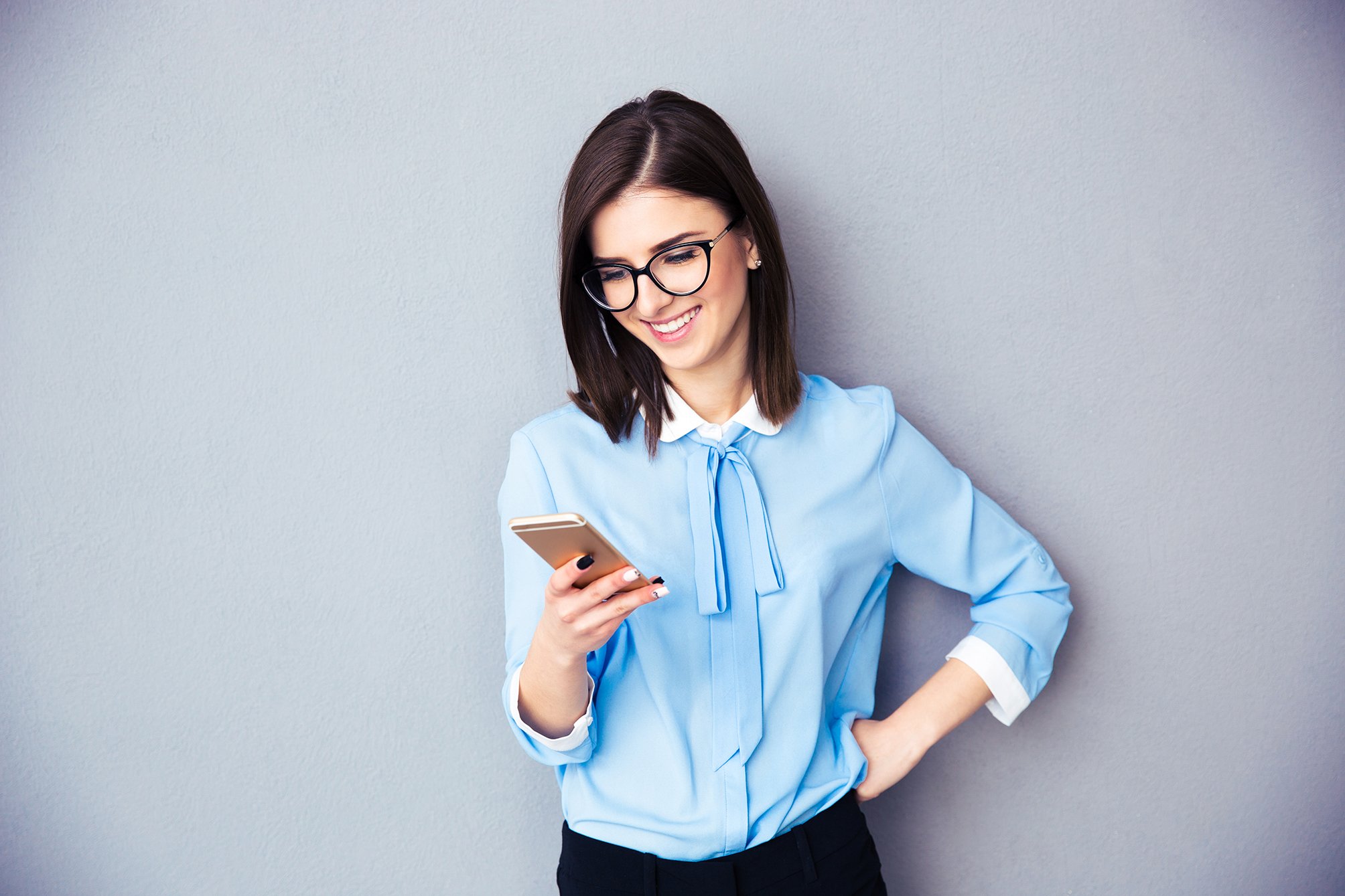 (150 dpi) smiling-businesswoman-using-smartphone-over-gray-background-wearing-in-blue-shirt-and-glasses